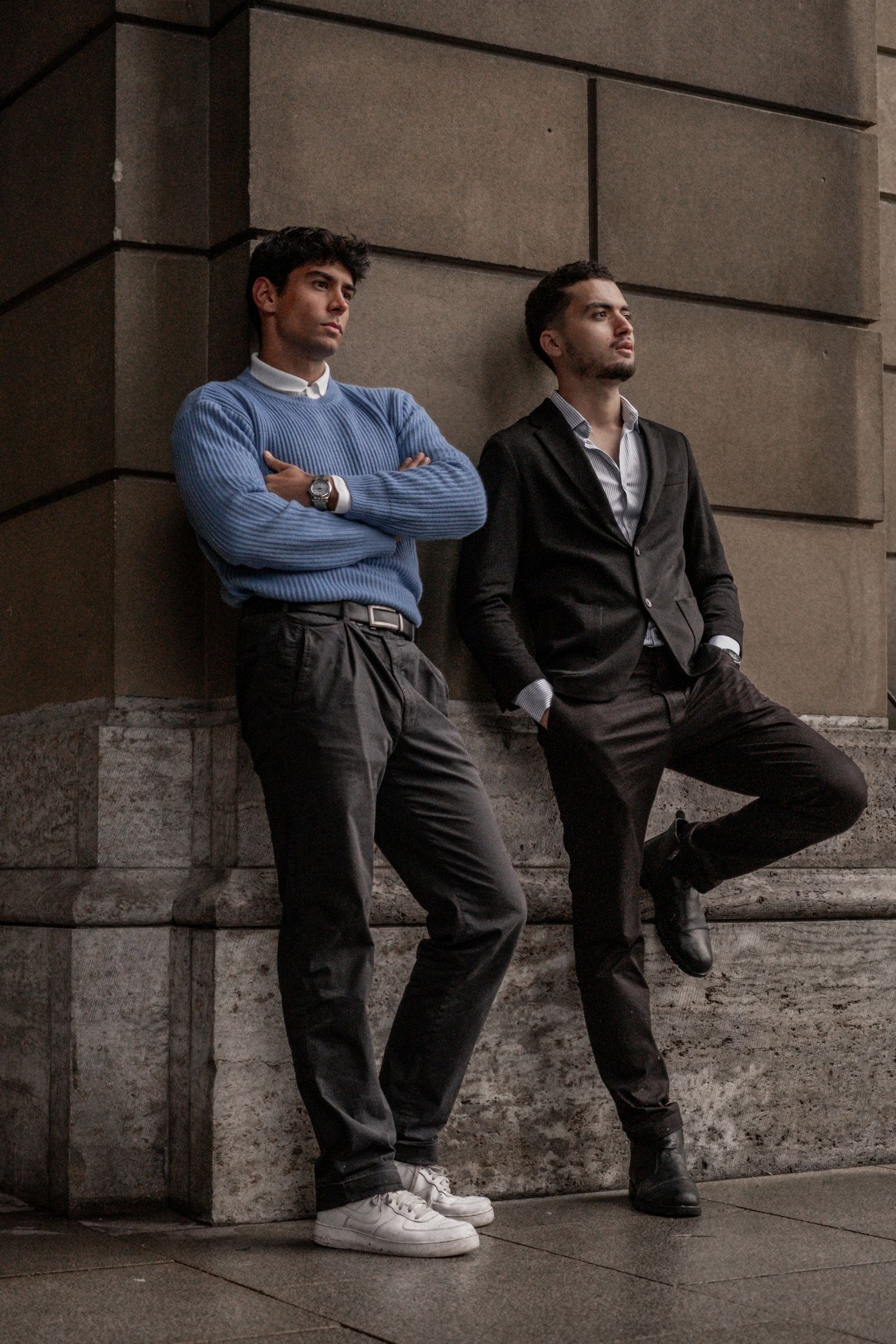 Two men in modern city outfits leaning against a wall